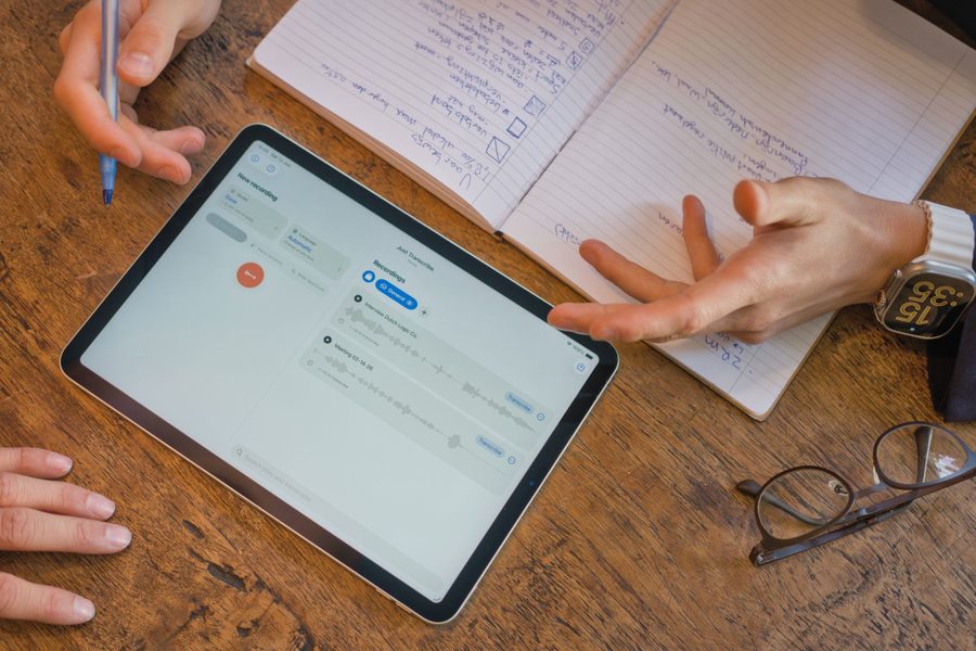 Un iPad su un tavolo di legno che mostra Just Transcribe in modalità chiara, accanto a un taccuino aperto con appunti scritti a mano.